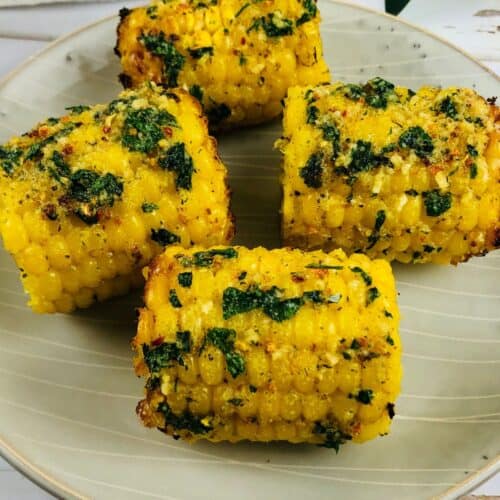 Four pieces of Air Fryer Corn on the cob, coated with herbs and spices, are arranged on a light-colored plate.