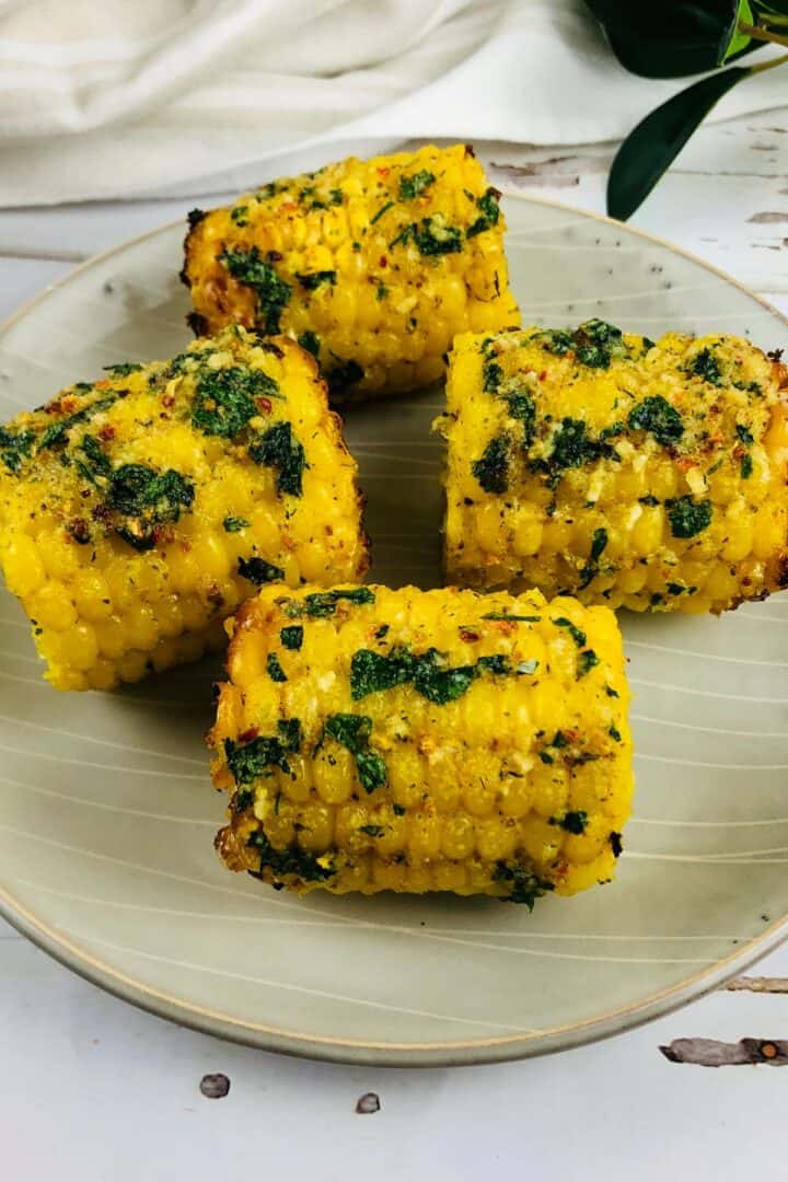 Four pieces of Air Fryer Corn on the cob, coated with herbs and spices, are arranged on a light-colored plate.