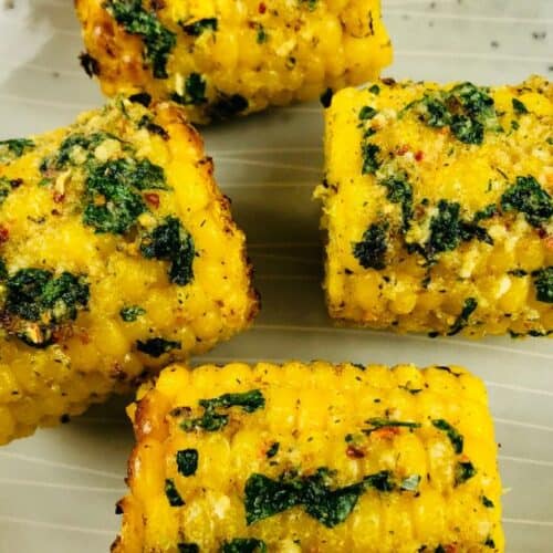 Four pieces of Air Fryer Corn on the cob, coated with herbs and spices, are arranged on a beige plate.