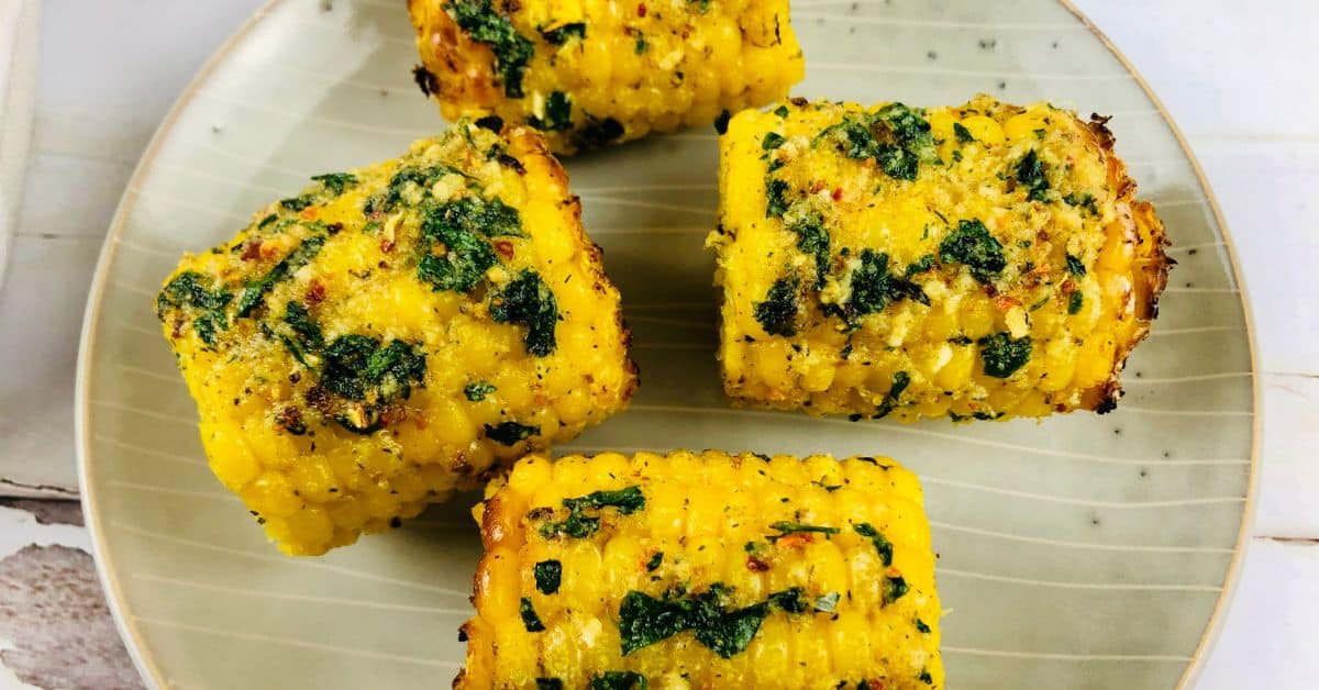 Four pieces of Air Fryer Corn on the cob, coated with herbs and spices, are arranged on a beige plate.