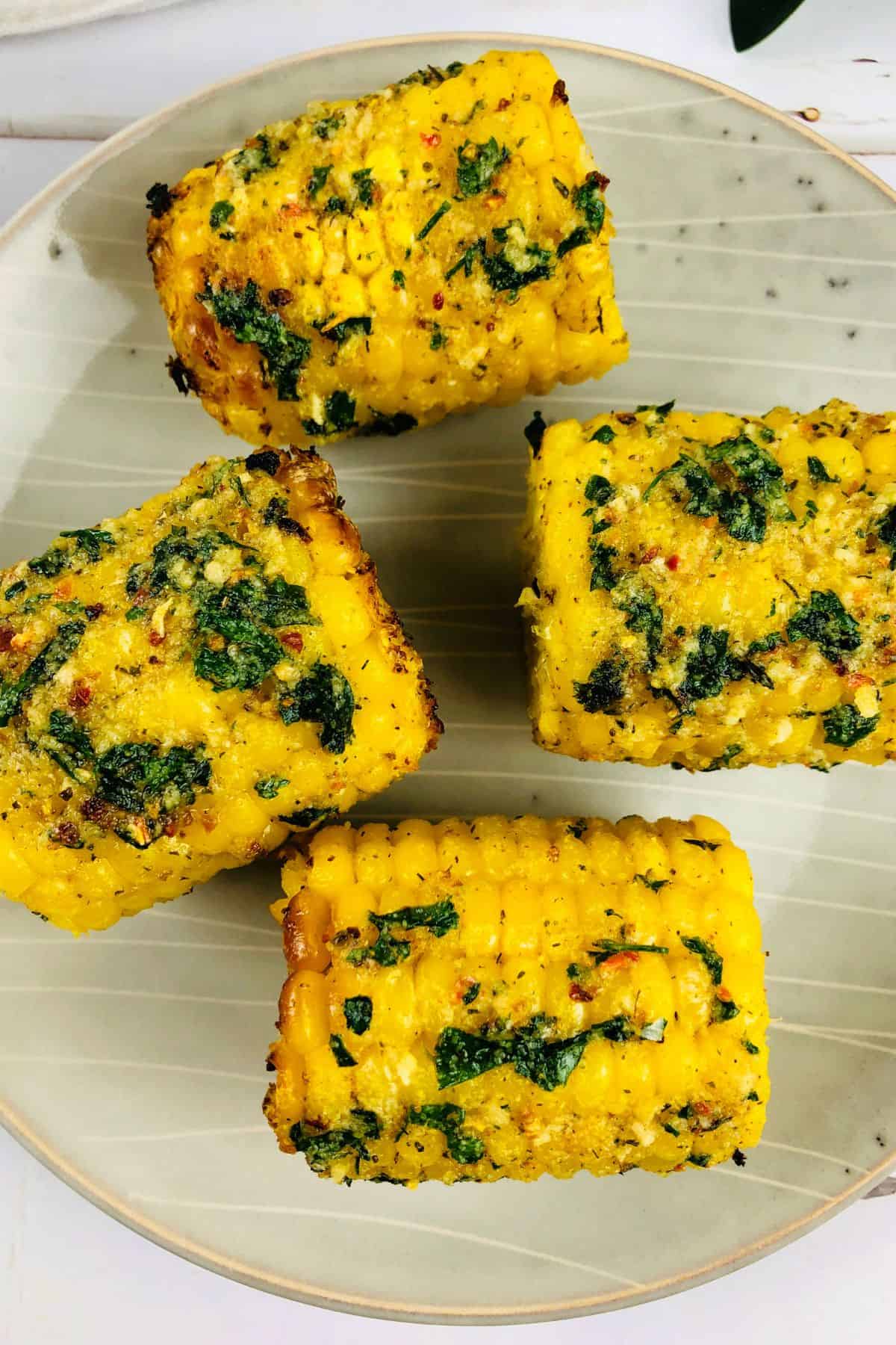Four pieces of Air Fryer Corn on the cob, covered with herbs and spices, are arranged on a beige plate.