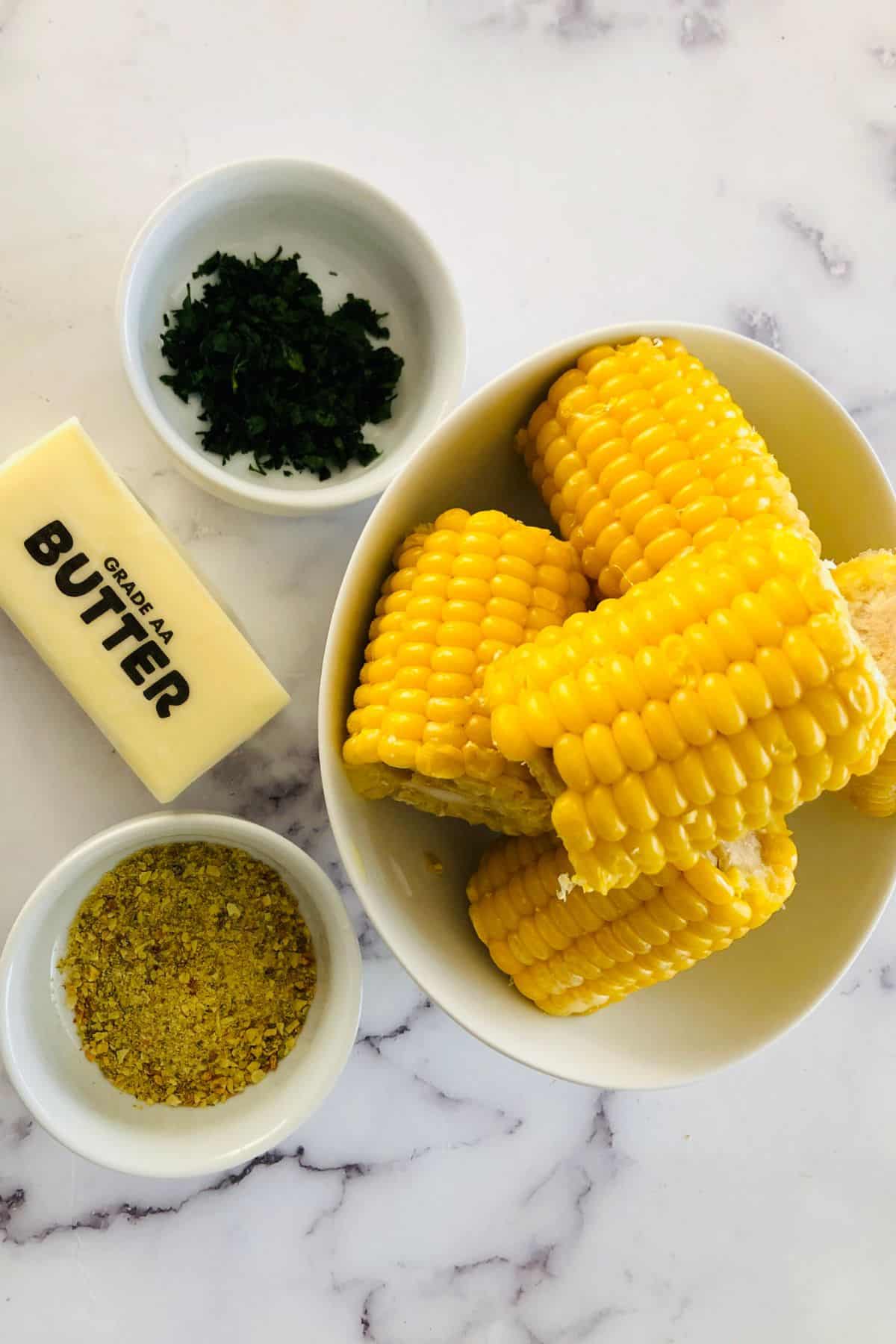 Bowl of Air Fryer Corn on the Cob with a stick of butter, chopped herbs in a small bowl, and a bowl of yellow seasoning on a white marble surface.