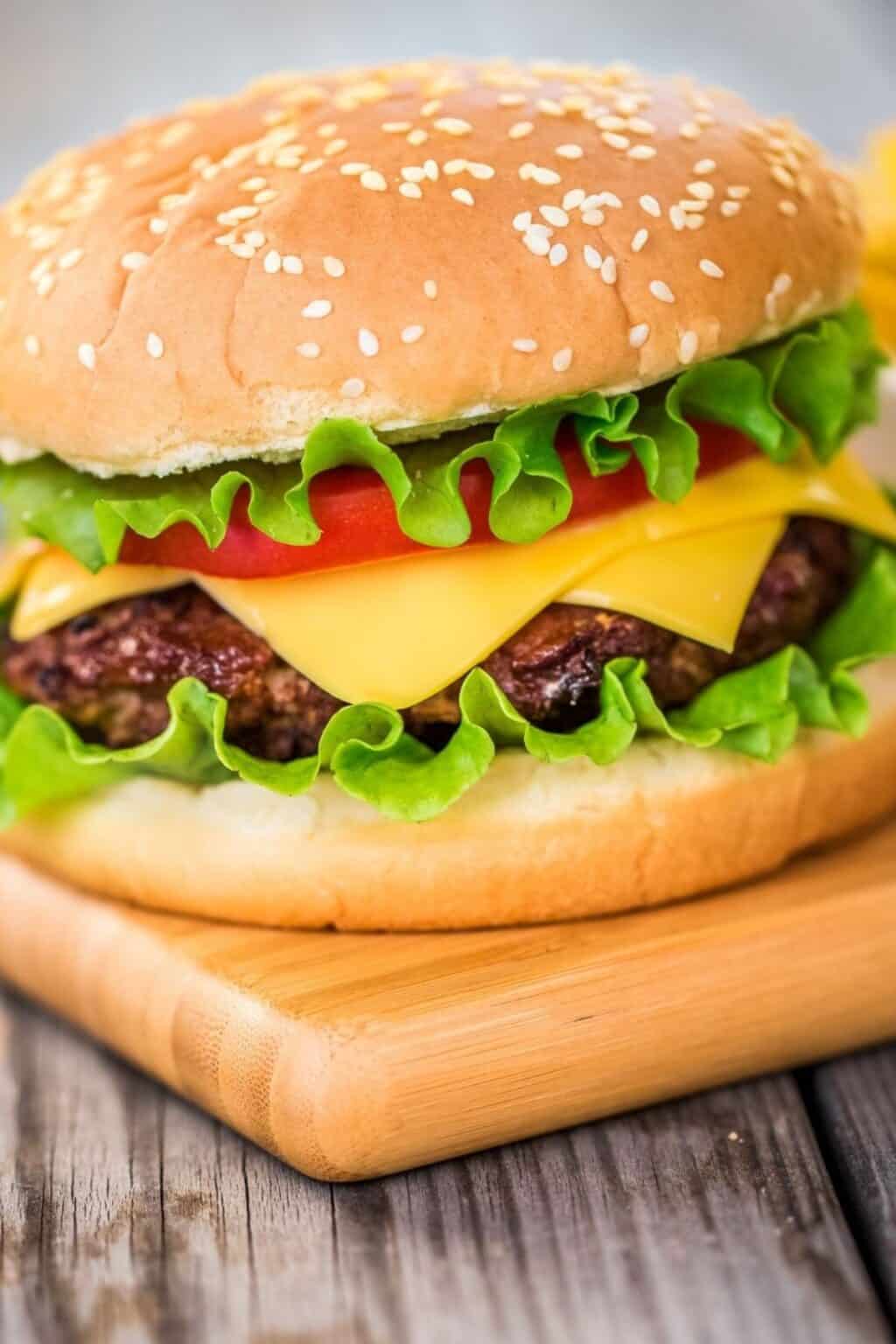 A cheeseburger with lettuce, tomato, and cheddar cheese in a sesame seed bun sits on a wooden board, showcasing a classic take on baked hamburgers.