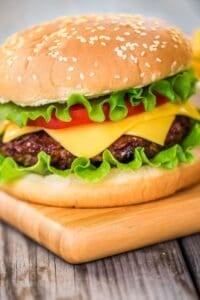 A cheeseburger with lettuce, tomato, and cheddar cheese in a sesame seed bun sits on a wooden board, showcasing a classic take on baked hamburgers.