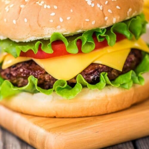 A cheeseburger with lettuce, tomato, and cheddar cheese in a sesame seed bun sits on a wooden board, showcasing a classic take on baked hamburgers.
