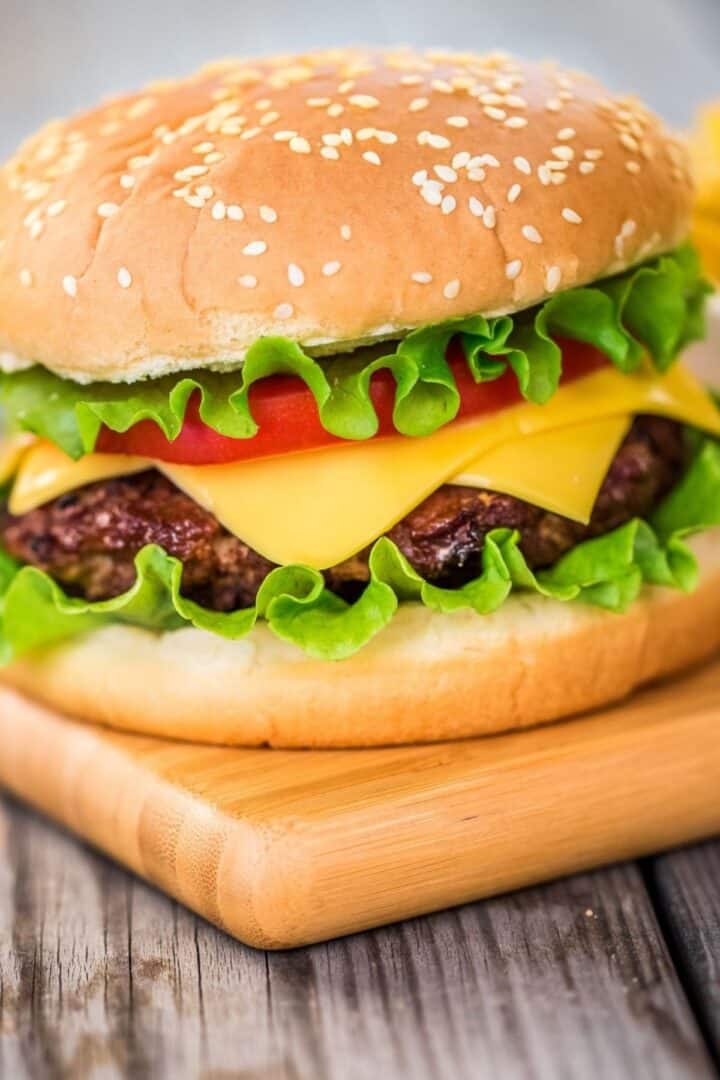 A cheeseburger with lettuce, tomato, and cheddar cheese in a sesame seed bun sits on a wooden board, showcasing a classic take on baked hamburgers.