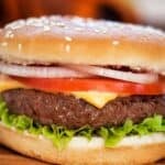 A cheeseburger with lettuce, tomato, onion, cheese, and a beef patty in a sesame seed bun sits on a wooden surface, showcasing the classic flavors of baked hamburgers.