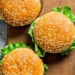 Three sesame seed hamburger buns filled with lettuce and cheese are arranged on a wooden cutting board, making these baked hamburgers an irresistible choice for your next meal.