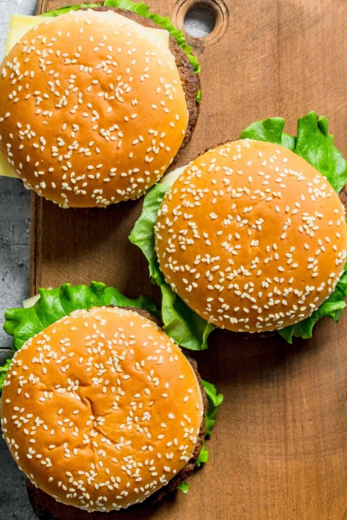 Three sesame seed hamburger buns filled with lettuce and cheese are arranged on a wooden cutting board, making these baked hamburgers an irresistible choice for your next meal.