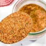 A close-up of a spoonful of reddish-brown burger seasoning mix with herbs, held above an open glass jar. A red checkered cloth is partially visible in the background.