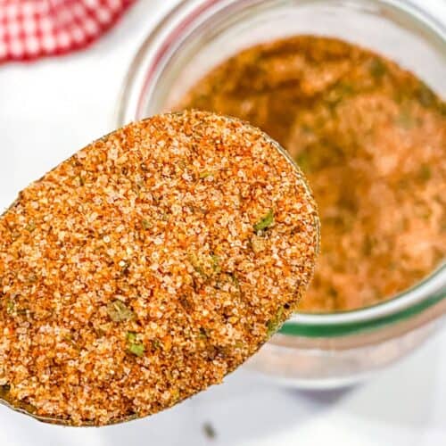 A close-up of a spoonful of reddish-brown burger seasoning mix with herbs, held above an open glass jar. A red checkered cloth is partially visible in the background.