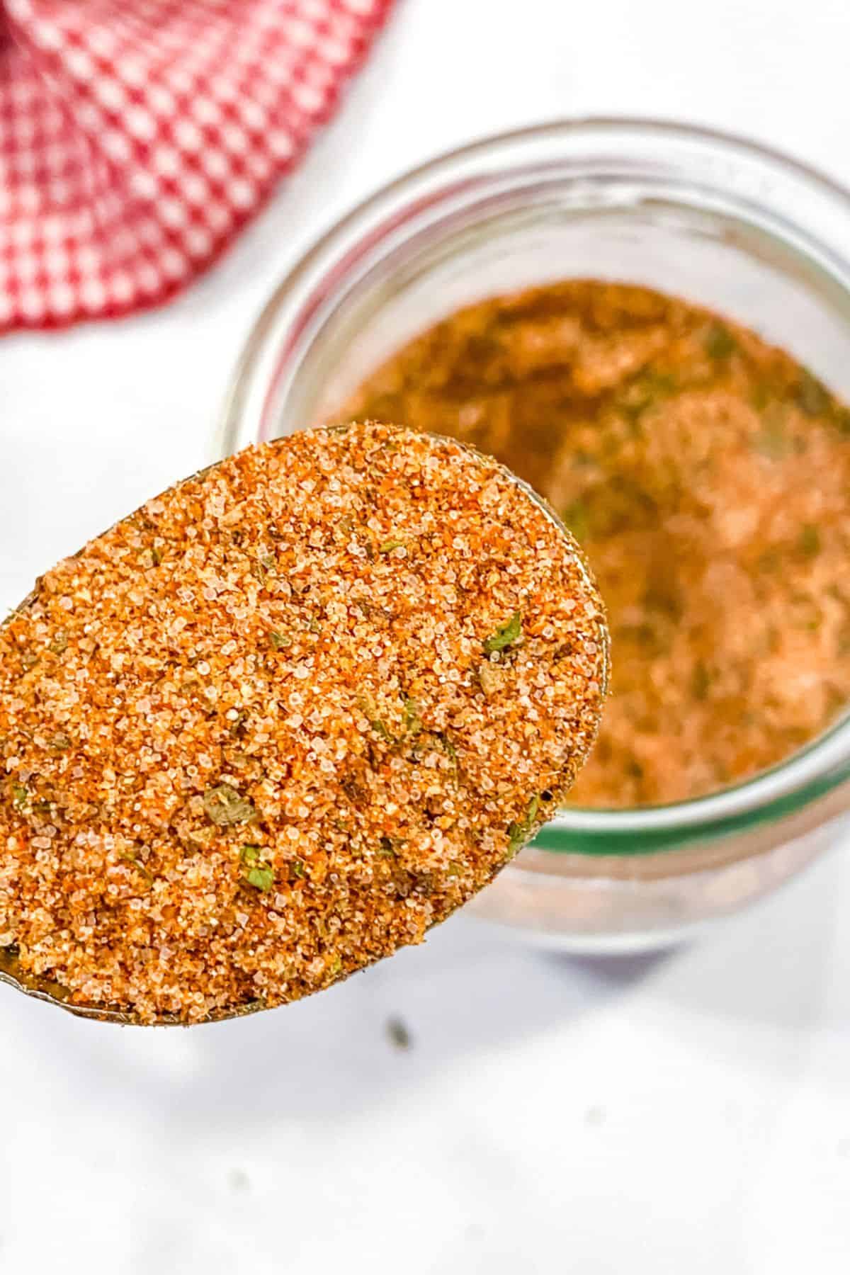 A close-up of a spoonful of reddish-brown burger seasoning mix with herbs, held above an open glass jar. A red checkered cloth is partially visible in the background.