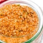 A spoonful of reddish-brown burger seasoning held over a glass jar, with a red and white checkered cloth in the background.