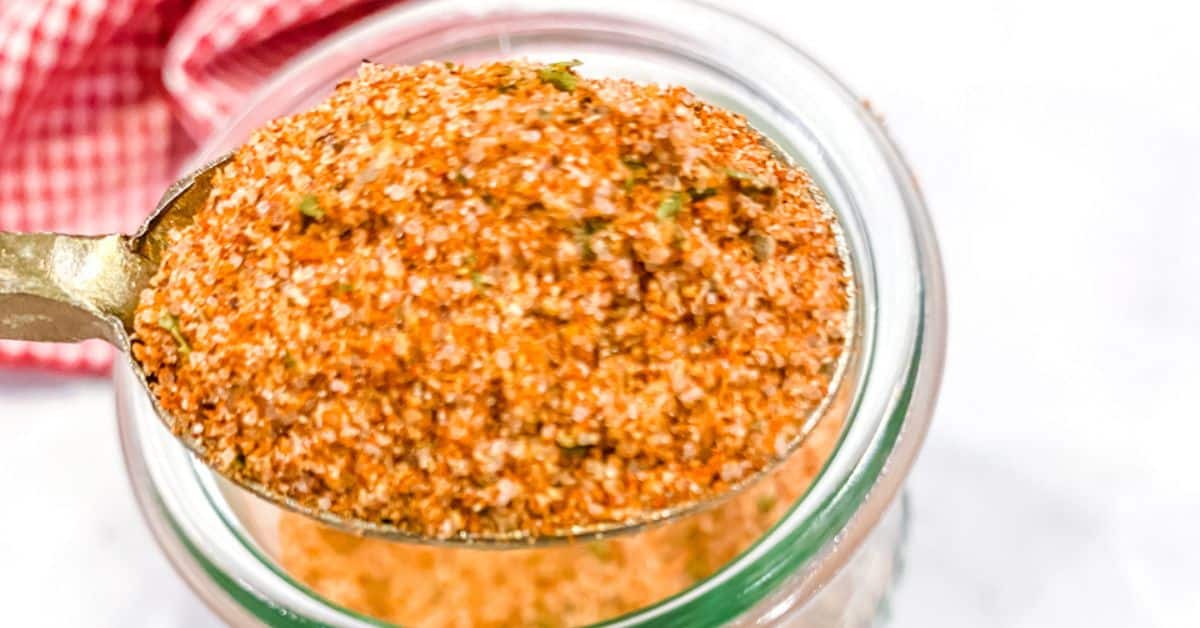 A spoonful of reddish-brown burger seasoning held over a glass jar, with a red and white checkered cloth in the background.