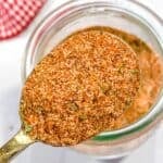 A spoon holds a blend of reddish-brown Burger Seasoning, with a jar of the same seasoning and a red checkered cloth in the background.