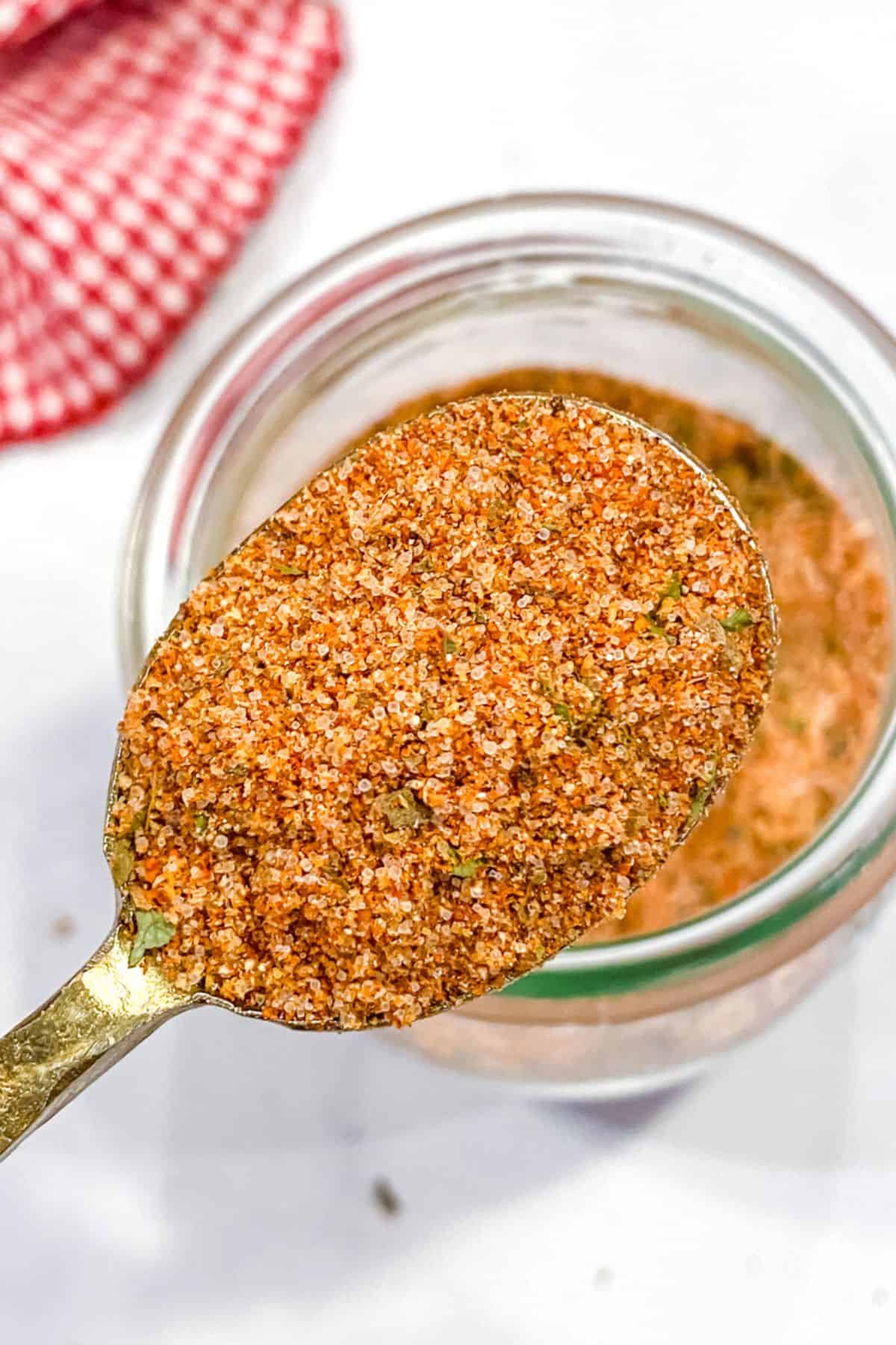 A spoon holds a blend of reddish-brown Burger Seasoning, with a jar of the same seasoning and a red checkered cloth in the background.