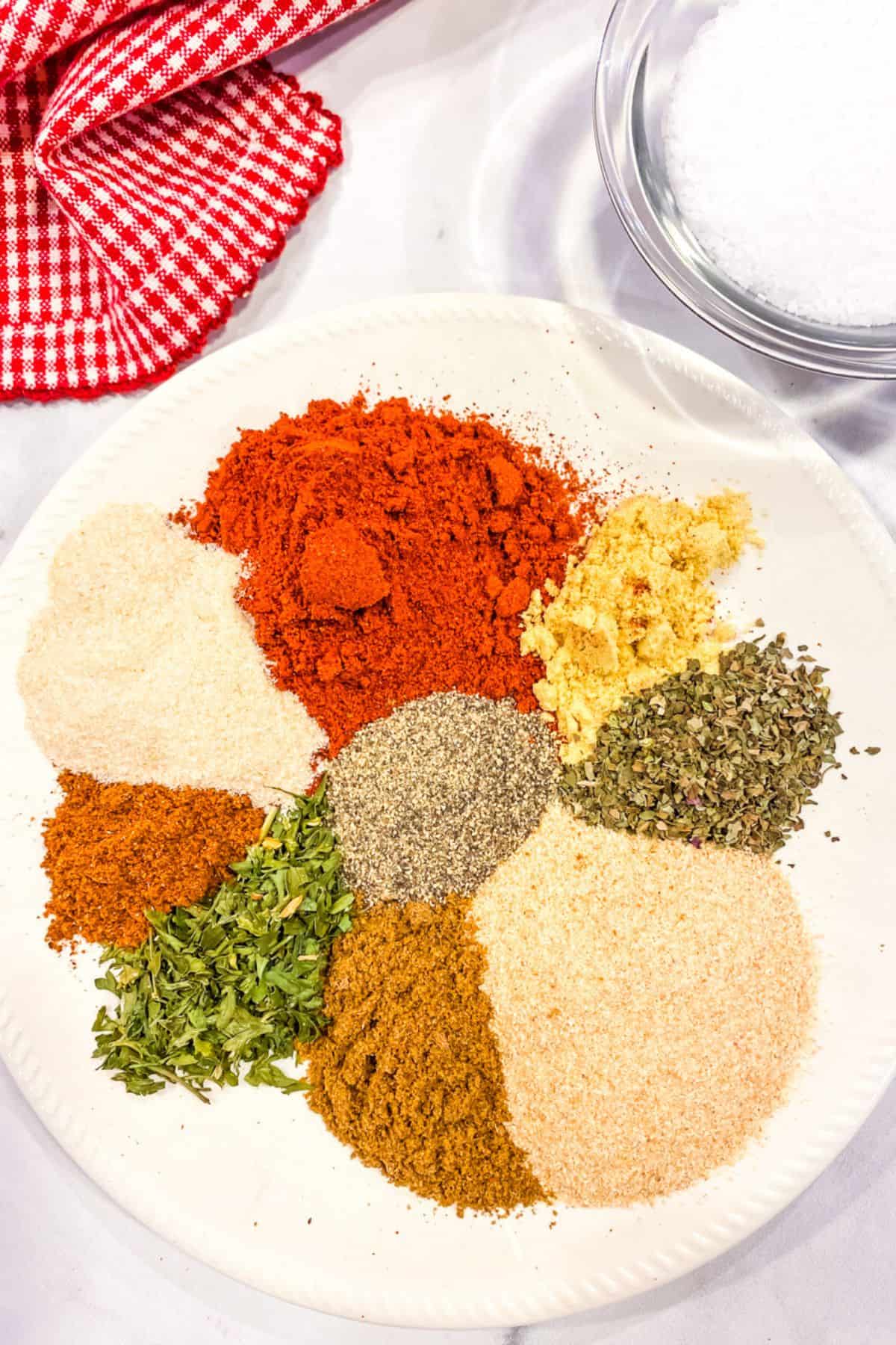 A white plate displays assorted powdered spices and herbs, including Burger Seasoning, arranged in small piles, with a red checkered cloth and a bowl of salt nearby.