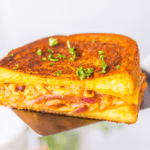 Close-up of a buffalo chicken grilled cheese sandwich with melted cheese and caramelized onions, garnished with chopped herbs, held on a metal spatula.