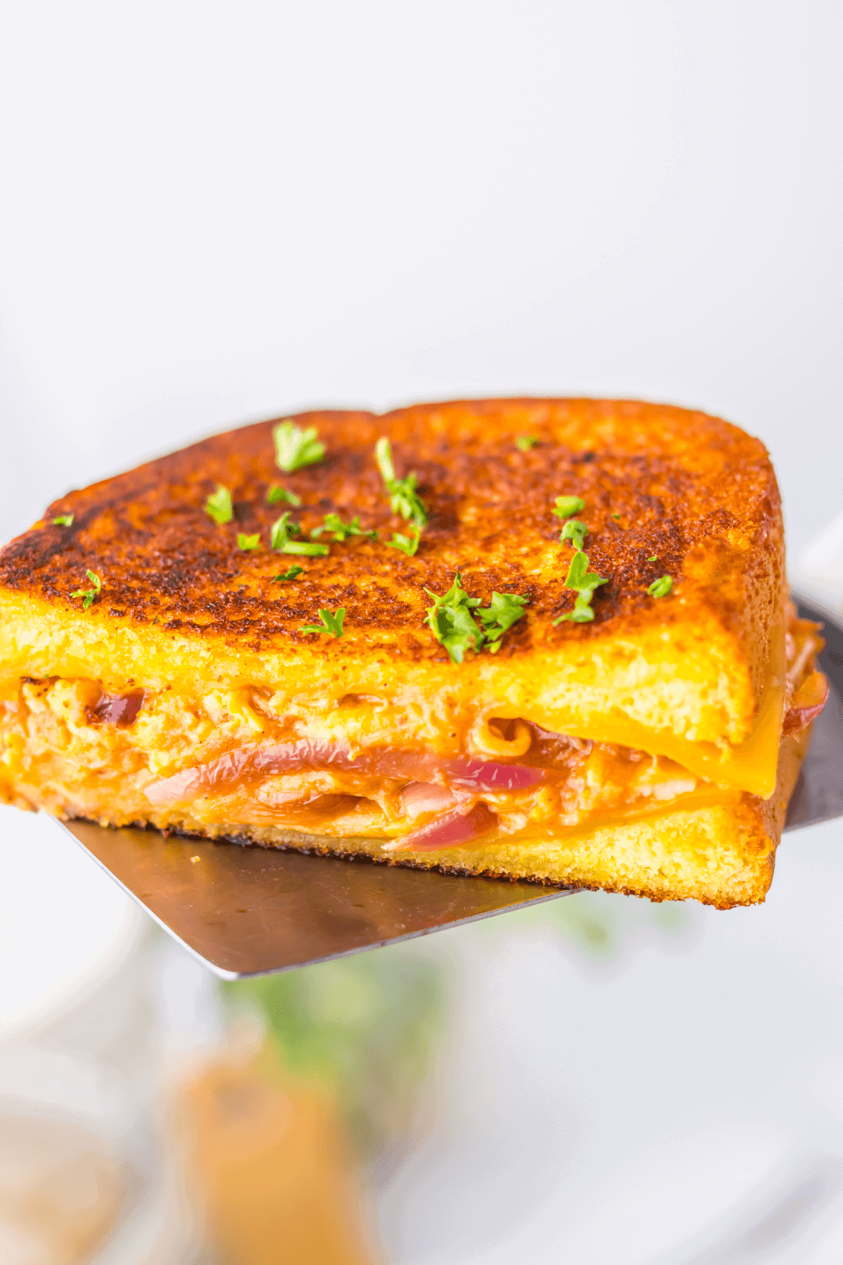 Close-up of a buffalo chicken grilled cheese sandwich with melted cheese and caramelized onions, garnished with chopped herbs, held on a metal spatula.