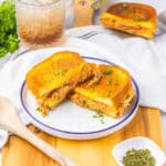 A plate with a cut grilled buffalo chicken grilled cheese sandwich, garnished with herbs. Nearby, there's a small dish of herbs, a wooden spoon, a drink with ice, and a leafy green plant.