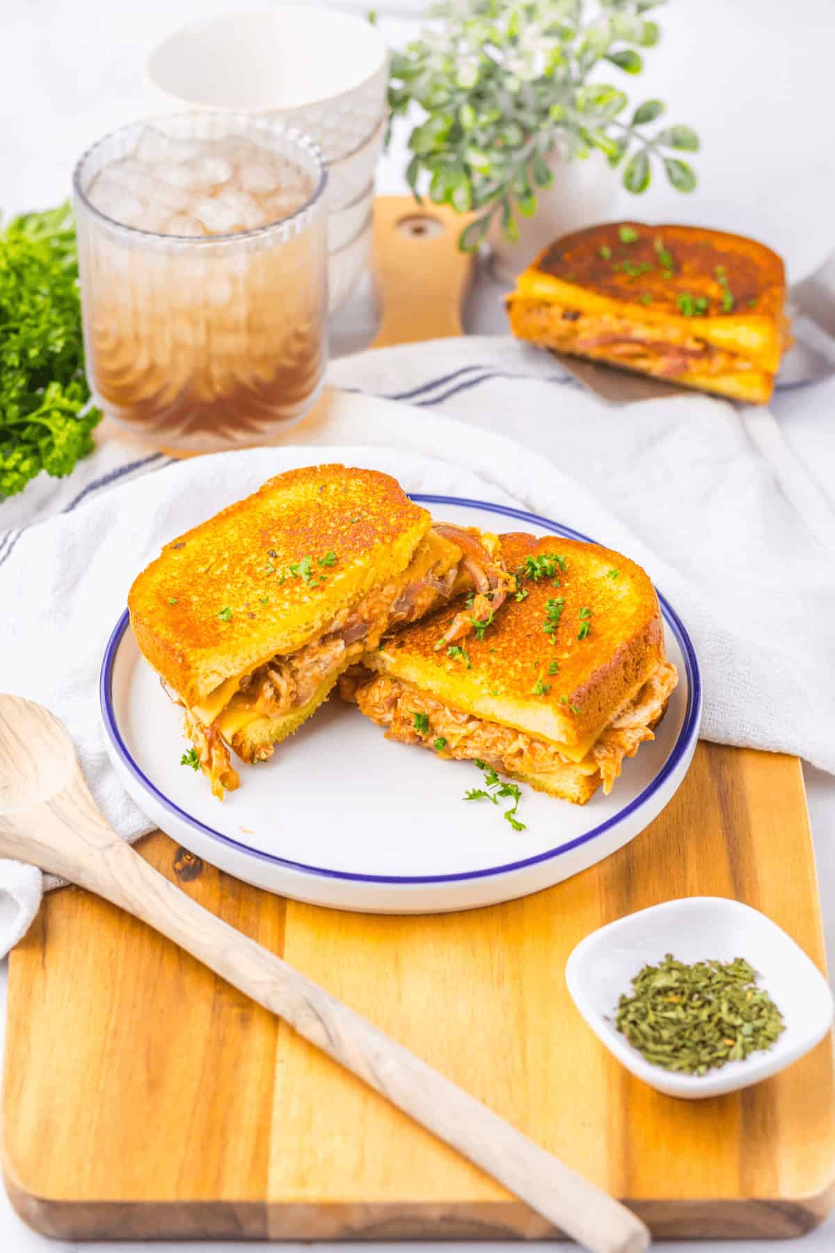 A plate with a cut grilled buffalo chicken grilled cheese sandwich, garnished with herbs. Nearby, there's a small dish of herbs, a wooden spoon, a drink with ice, and a leafy green plant.