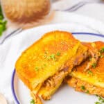 A buffalo chicken grilled cheese sandwich, lavishly stuffed with shredded meat, is cut in half and placed on a white plate with a blue rim. In the background, a glass of iced drink complements the meal perfectly.