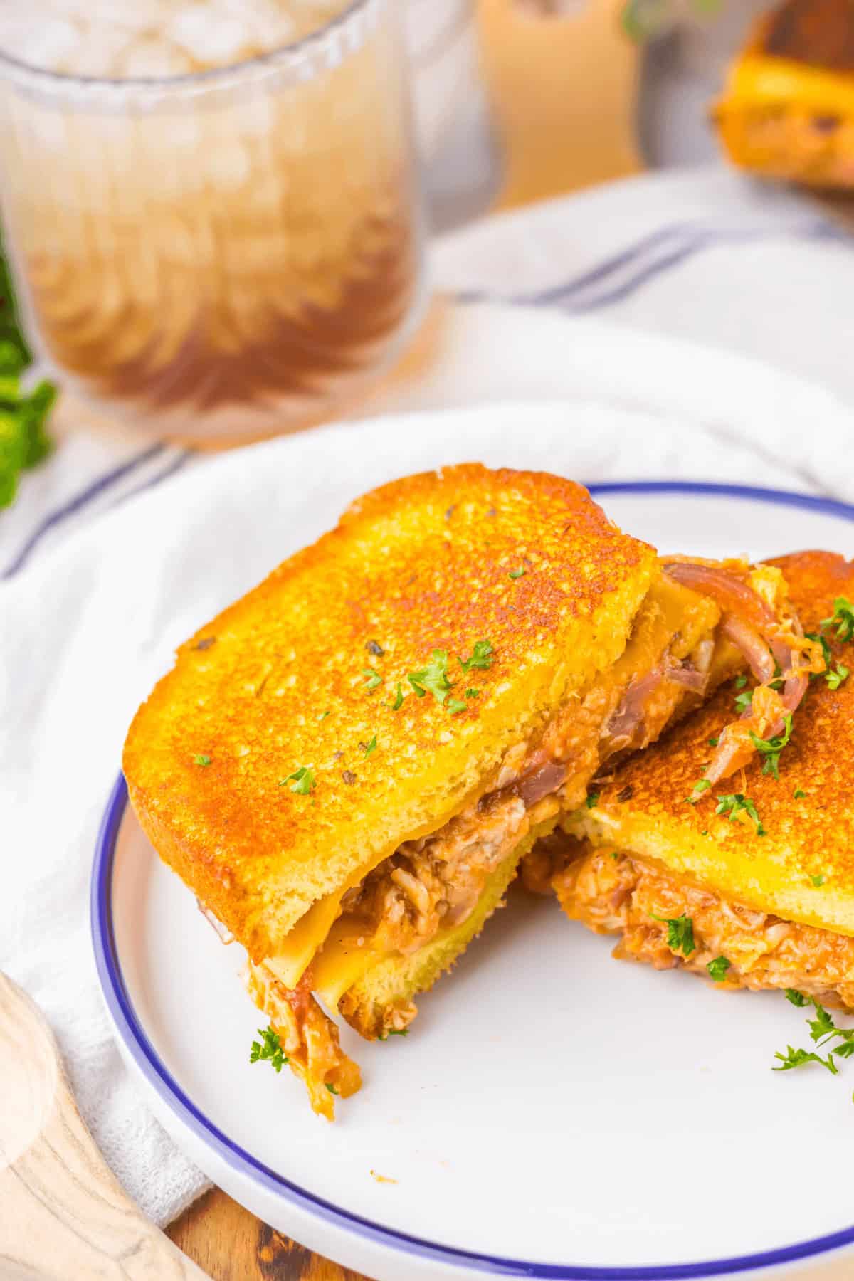 A buffalo chicken grilled cheese sandwich, lavishly stuffed with shredded meat, is cut in half and placed on a white plate with a blue rim. In the background, a glass of iced drink complements the meal perfectly.