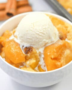 A white bowl filled with peach cobbler with cake mix topped with a scoop of vanilla ice cream.