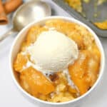 A bowl of peach cobbler with cake mix topped with a scoop of vanilla ice cream, with cinnamon sticks and a metal spoon nearby.
