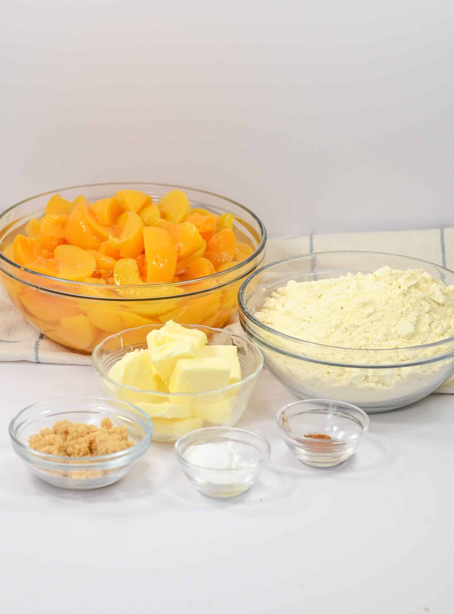 Ingredients for a peach cobbler with cake mix arranged on a white surface, including bowls of peach slices, flour, butter, brown sugar, baking powder, and ground cinnamon.