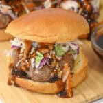 A close-up of a pork burger with a glossy bun, topped with coleslaw and drizzled with a dark sauce, placed on a wooden surface.