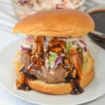 A pork burger on a plate topped with shredded lettuce, coleslaw, and drizzled with barbecue sauce. A bowl of coleslaw and a dish of barbecue sauce are in the background.