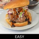 A pork burger topped with coleslaw and sauce sits on a white plate, with a bowl of coleslaw and a small dish of sauce in the background. Text below the image reads "Easy Pork Burgers." These delectable pork burgers are perfect for any meal.