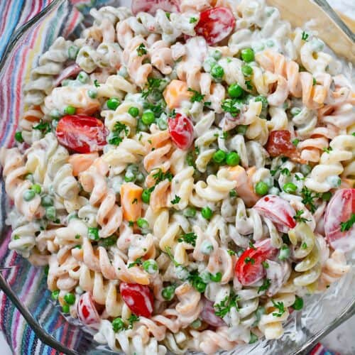 A glass bowl filled with creamy rotini pasta salad, mixed with cherry tomatoes, peas, diced cheese, and chopped herbs—this Bacon Pasta Salad with Ranch Dressing is beautifully displayed on a colorful striped cloth.