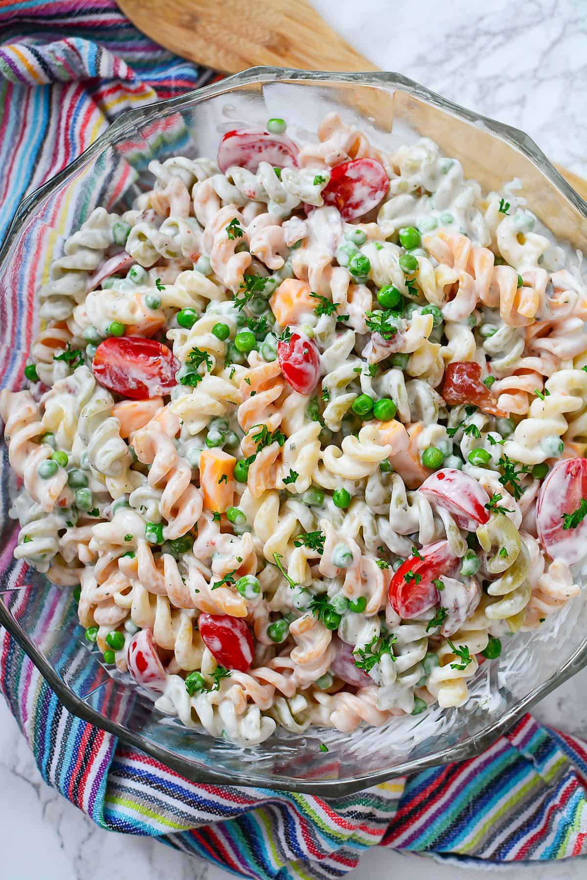 A glass bowl filled with creamy rotini pasta salad, mixed with cherry tomatoes, peas, diced cheese, and chopped herbs—this Bacon Pasta Salad with Ranch Dressing is beautifully displayed on a colorful striped cloth.