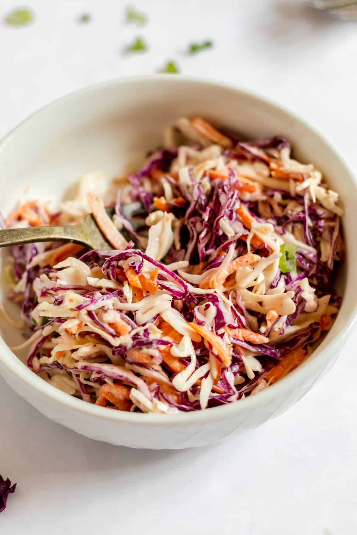 A white bowl filled with Southern Coleslaw made of shredded red and green cabbage and carrots, mixed with a creamy dressing, with a fork inside.