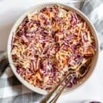 A bowl of Southern Coleslaw with shredded red and green cabbage and carrots, mixed with dressing, sits ready to serve with two spoons on a checkered cloth.