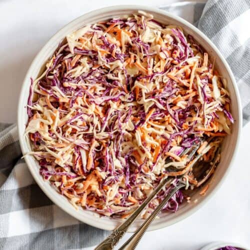 A bowl of Southern Coleslaw with shredded red and green cabbage and carrots, mixed with dressing, sits ready to serve with two spoons on a checkered cloth.