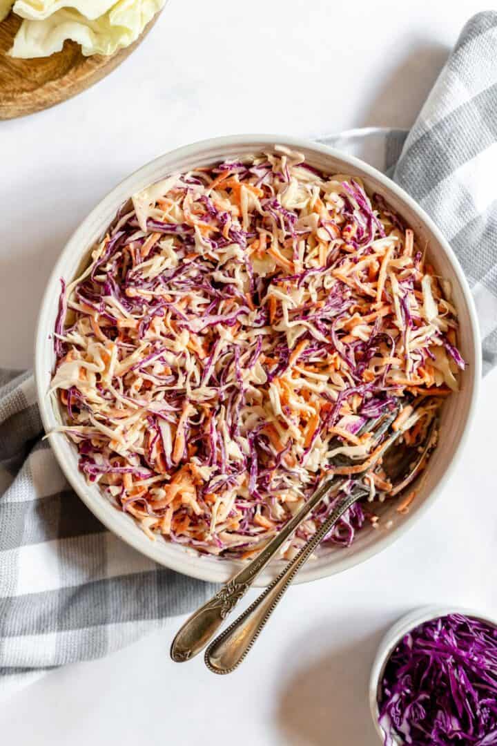 A bowl of Southern Coleslaw with shredded red and green cabbage and carrots, mixed with dressing, sits ready to serve with two spoons on a checkered cloth.