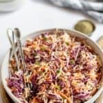 A bowl of Southern Coleslaw made with shredded green and purple cabbage and carrots, garnished with sliced green onions, with serving utensils and a small dish of pepper nearby.