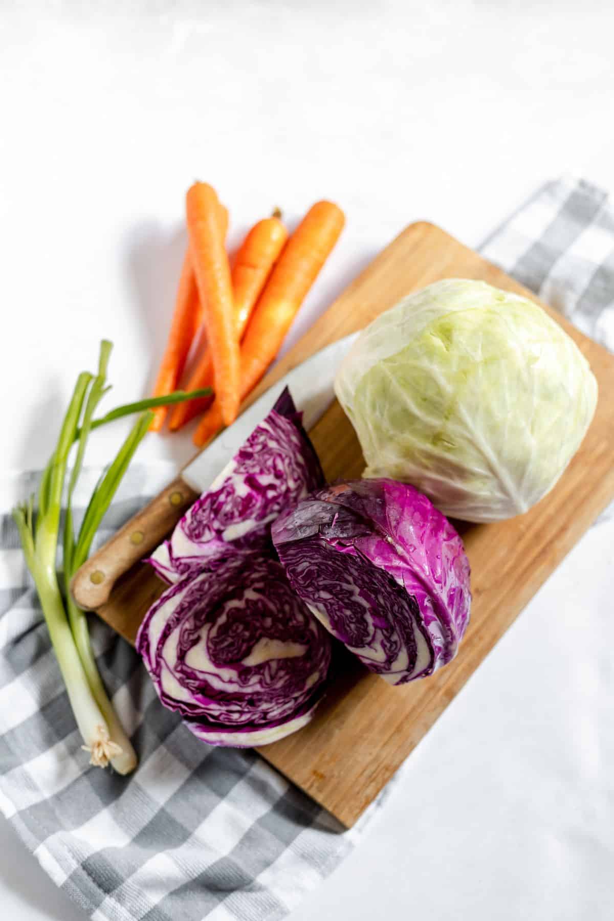 A cutting board with a whole green cabbage, sliced red cabbage, carrots, and green onions on a checkered cloth. A knife rests beside the vegetables.