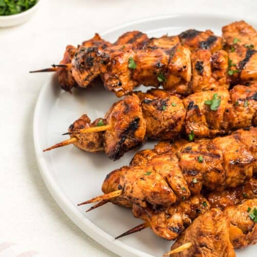 Grilled chicken on a stick is artfully arranged on a white plate and garnished with fresh herbs. In the background, a bowl of savory sauce and a small dish of chopped herbs complete the inviting presentation.