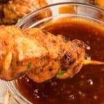 A delicious grilled chicken on a stick is being dipped into a bowl of reddish-brown sauce.