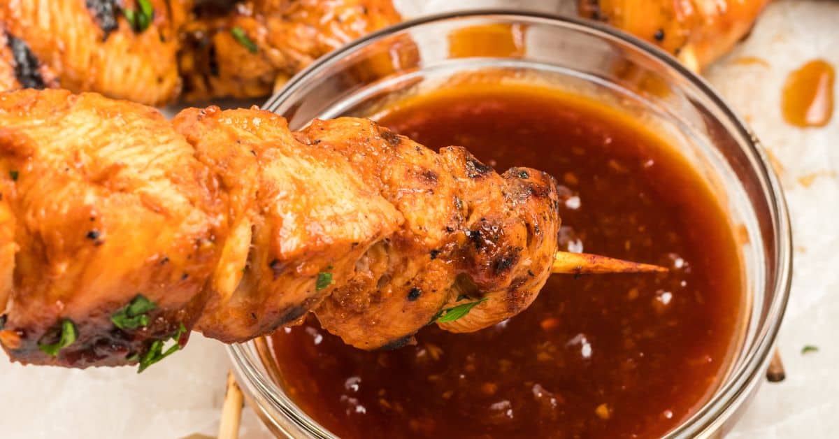 A delicious grilled chicken on a stick is being dipped into a bowl of reddish-brown sauce.
