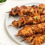 Skewers are arranged on a white plate, garnished with fresh herbs, with a small bowl of sauce and a dish of chopped herbs in the background.