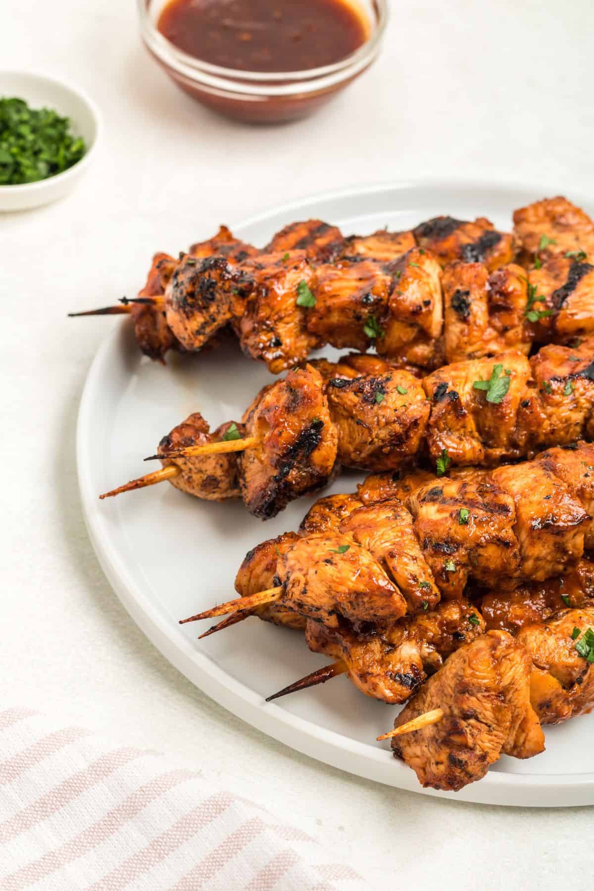 Skewers are arranged on a white plate, garnished with fresh herbs, with a small bowl of sauce and a dish of chopped herbs in the background.