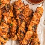 Grilled chicken on a stick with a reddish marinade are arranged on parchment paper next to a bowl of dipping sauce.