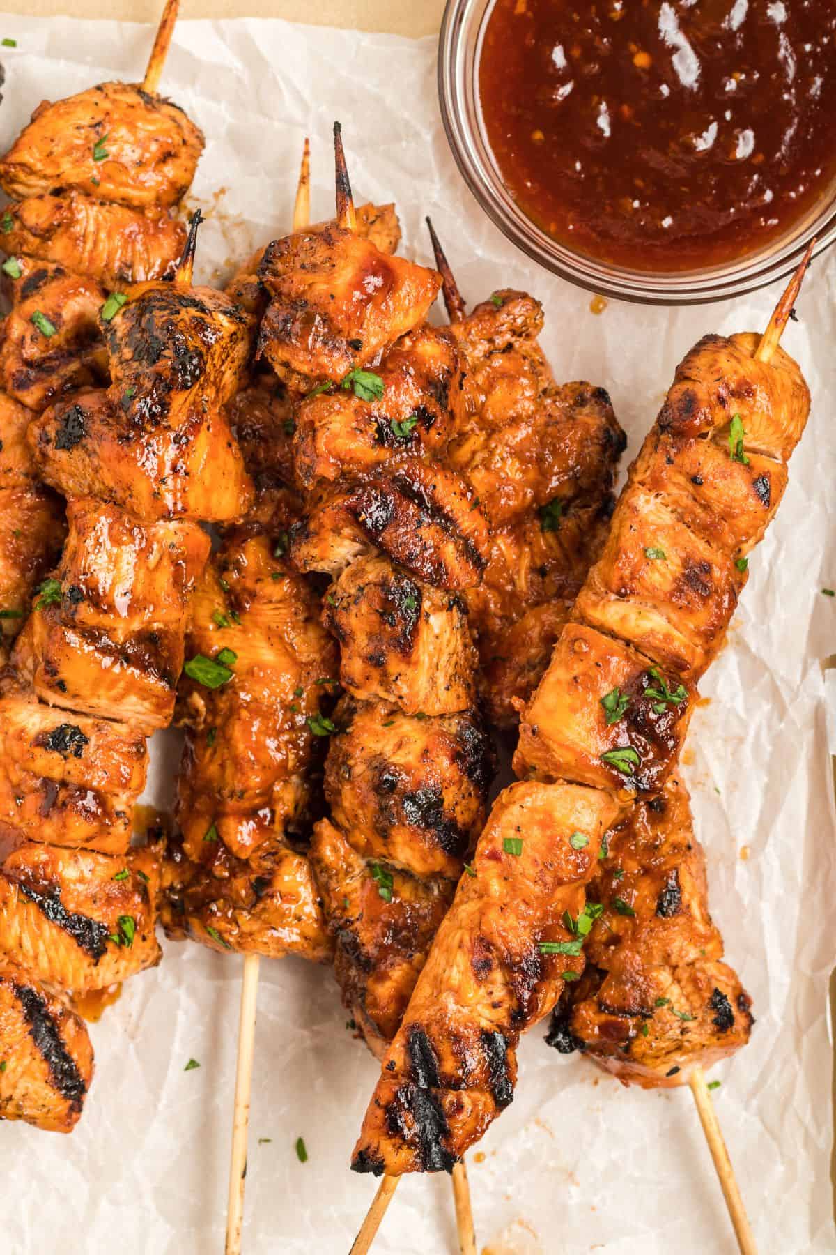 Grilled chicken on a stick with a reddish marinade are arranged on parchment paper next to a bowl of dipping sauce.