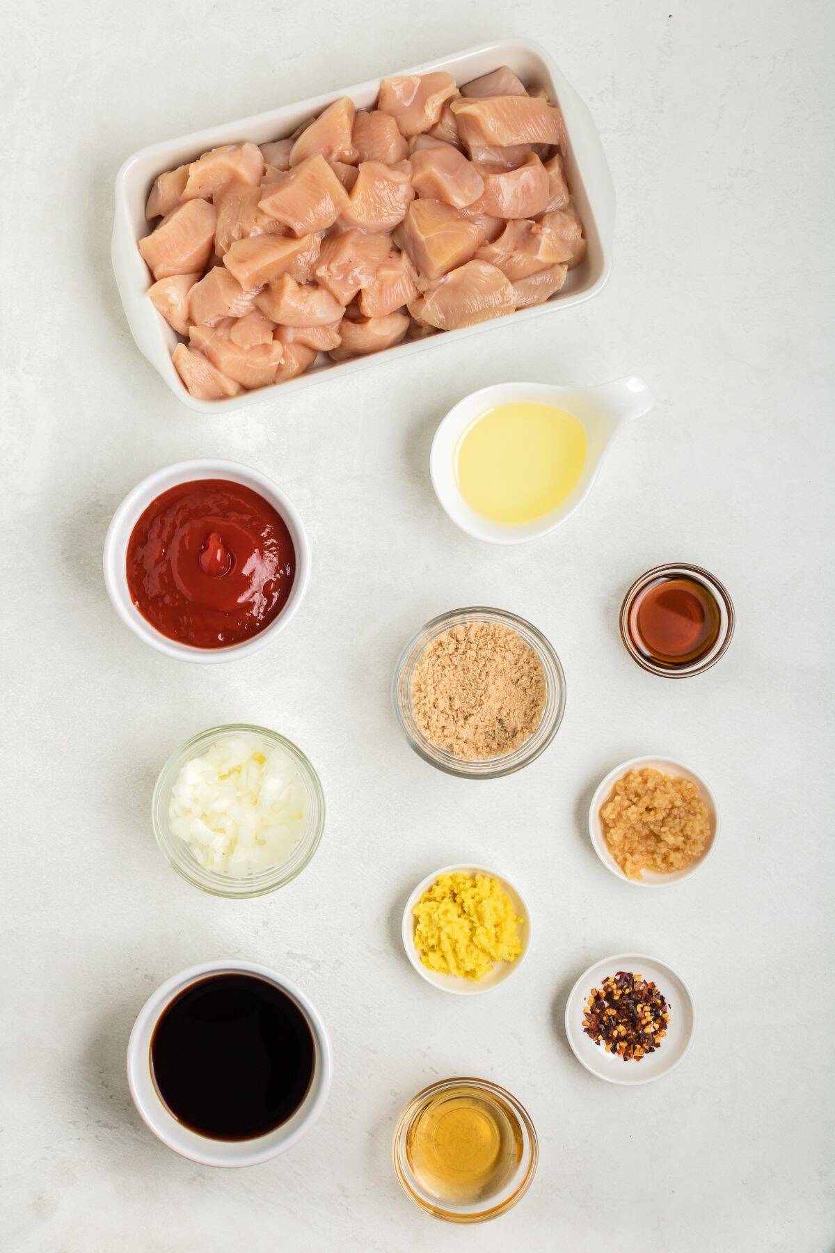 Overhead view of raw diced chicken in a dish, perfect for making chicken on a stick, surrounded by small bowls containing various sauces, liquids, minced garlic, chopped onion, spices, and breadcrumbs on a white surface.