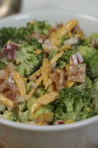 Close-up of an Amish broccoli salad with shredded cheddar cheese, red onion, bacon pieces, and a creamy dressing in a white bowl.